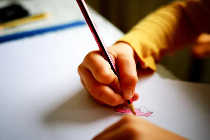 A child's hand drawing a pink house with a coloured pencil